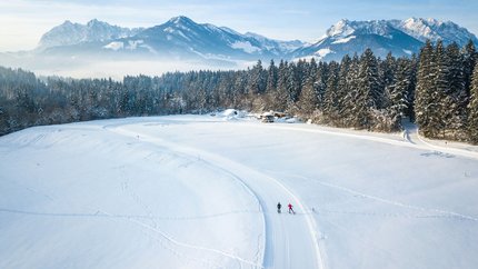 Two cross-country skiers on snowy trail with mountains and forest background