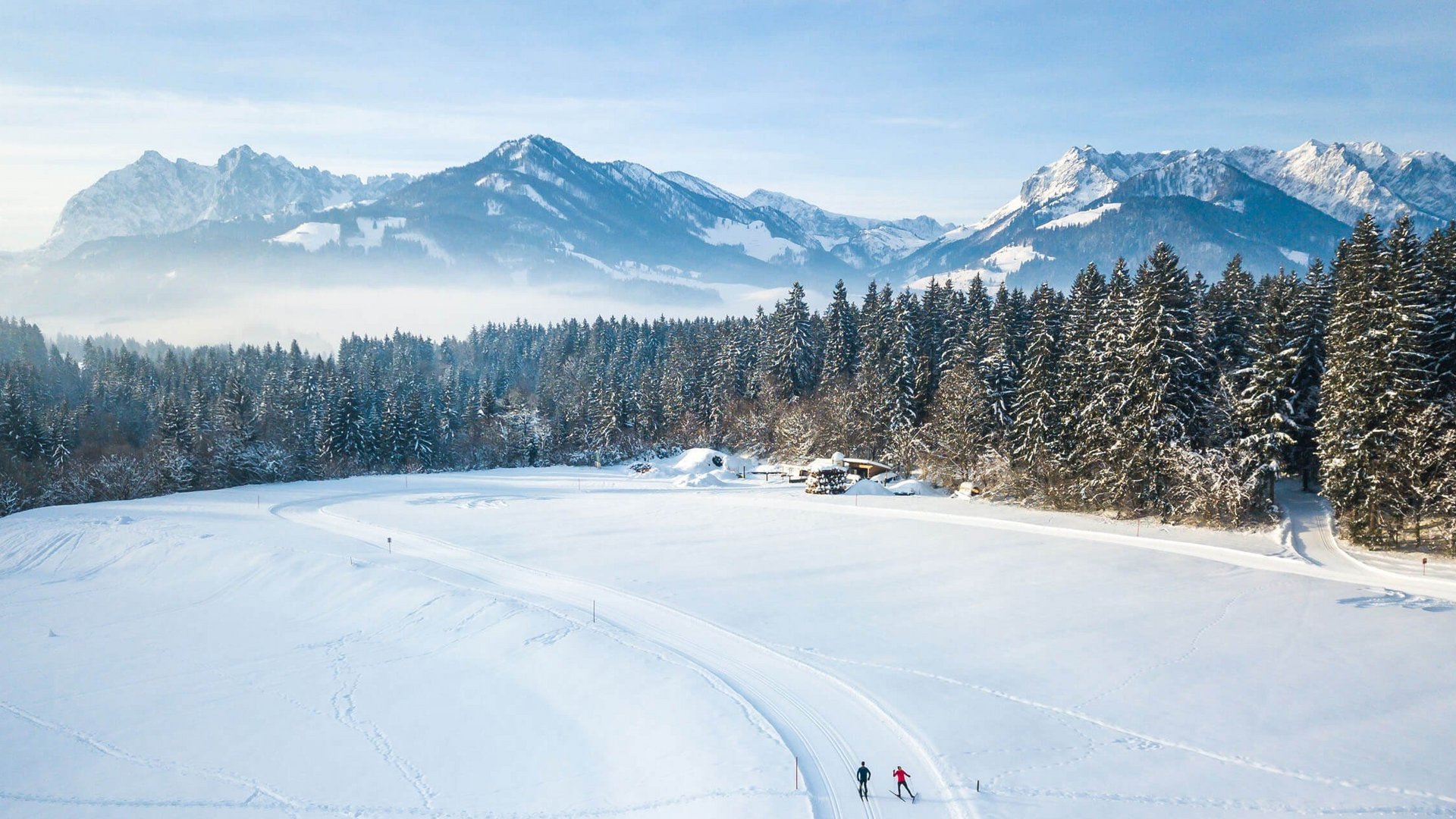Two cross-country skiers on snowy trail with mountains and forest background