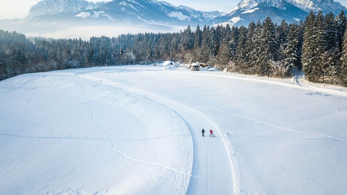 Kaiserwinkl: eine Langlaufregion der Superlative © Tourismusverband Kaiserwinkl Zwei Skifahrer auf verschneiter Loipe vor verschneiten Bergen und Wald