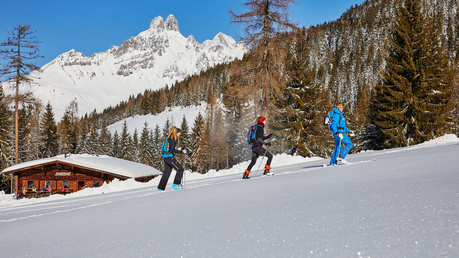 Drei Personen beim Schneeschuhwandern vor verschneiten Bergen und Hütte