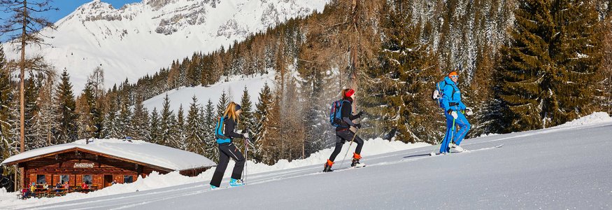 Drei Personen beim Schneeschuhwandern vor verschneiten Bergen und Hütte