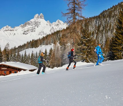 Drei Personen beim Schneeschuhwandern vor verschneiten Bergen und Hütte