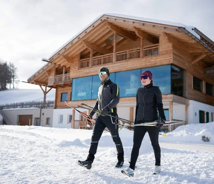 Zwei Skifahrer tragen Langlaufskier vor einem Chalet im Schnee