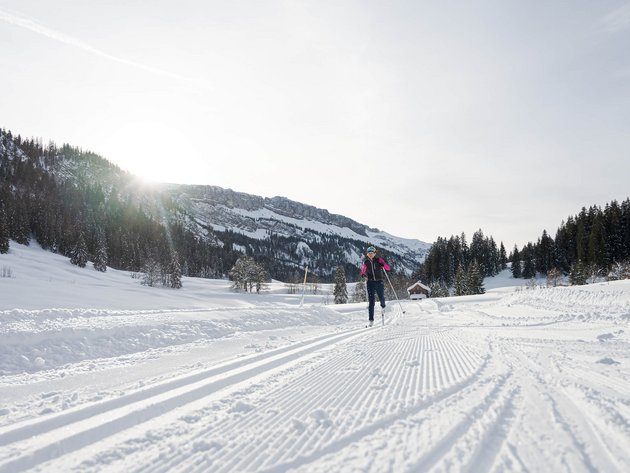 Person beim Skilanglauf auf verschneiter Loipe vor Bergkulisse bei Sonnenschein
