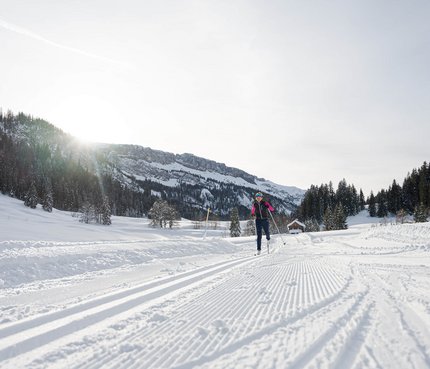 Person beim Skilanglauf auf verschneiter Loipe vor Bergkulisse bei Sonnenschein