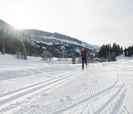 Person beim Skilanglauf auf verschneiter Loipe vor Bergkulisse bei Sonnenschein