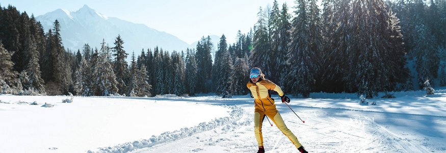 Langläuferin in gelber Kleidung auf schneebedeckter Loipe vor Tannen und Bergen