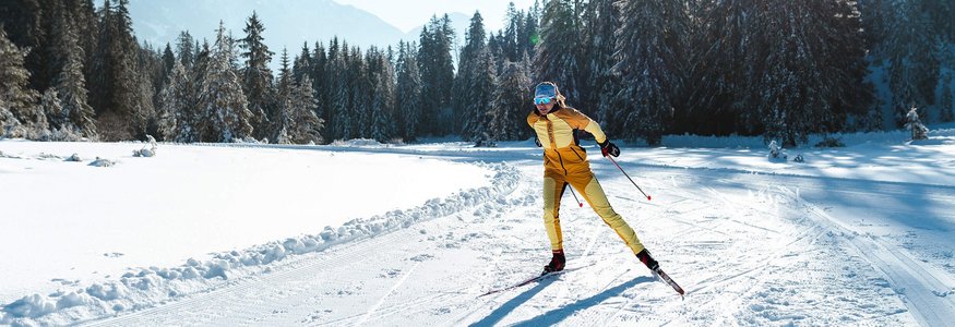 Langläuferin in gelber Kleidung auf schneebedeckter Loipe vor Tannen und Bergen