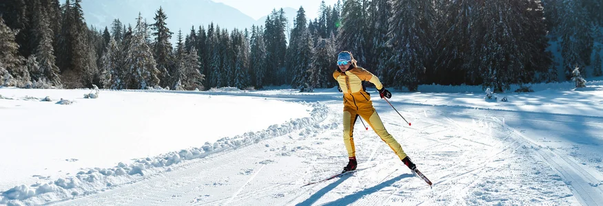 Langläuferin in gelber Kleidung auf schneebedeckter Loipe vor Tannen und Bergen