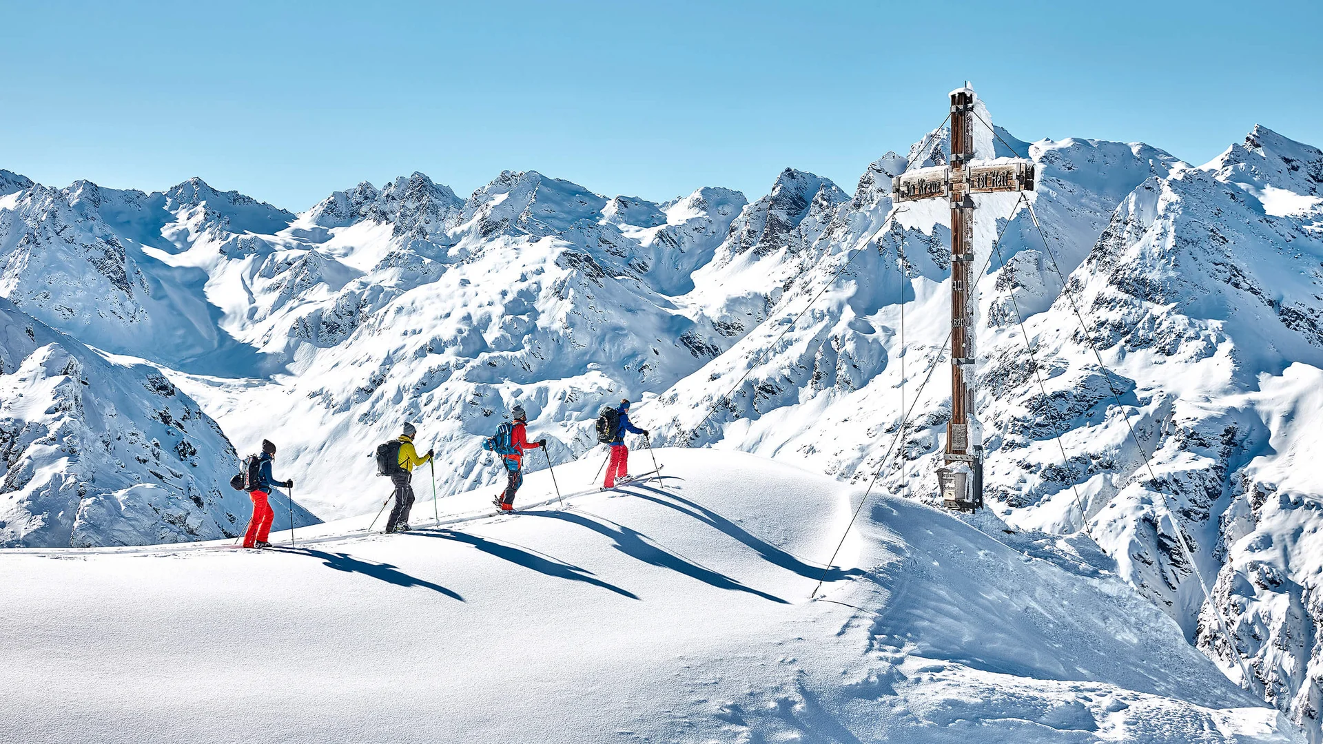 Vier Skifahrer auf schneebedecktem Gipfel mit großem Gipfelkreuz