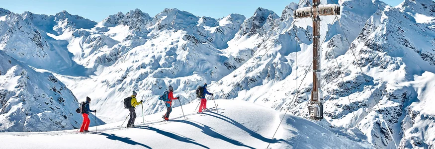 Vier Skifahrer auf schneebedecktem Gipfel mit großem Gipfelkreuz