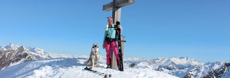 Skifahrerin mit Husky vor großem Holzkreuz auf schneebedecktem Berggipfel