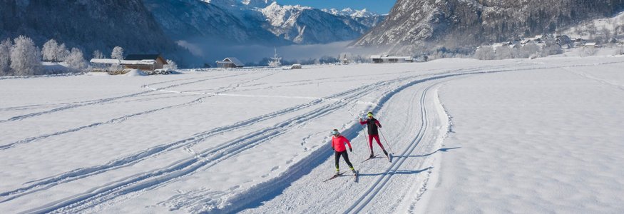 Zwei Langläufer auf schneebedeckter Loipe vor Bergen bei blauem Himmel