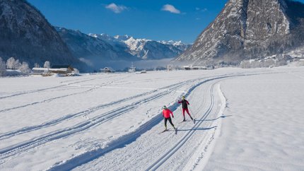 Two cross-country skiers on snowy trail with mountains and blue sky
