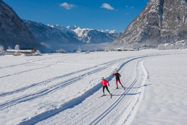 Two cross-country skiers on snowy trail with mountains and blue sky