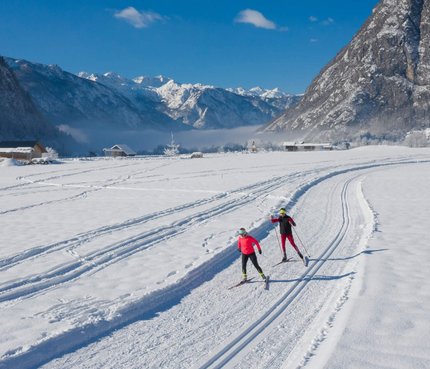 Zwei Langläufer auf schneebedeckter Loipe vor Bergen bei blauem Himmel