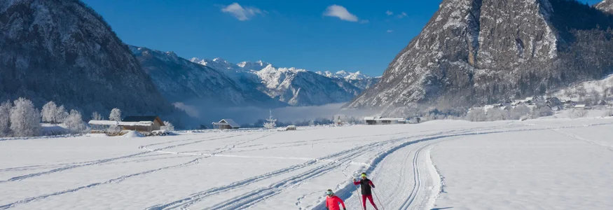 Zwei Langläufer auf schneebedeckter Loipe vor Bergen bei blauem Himmel