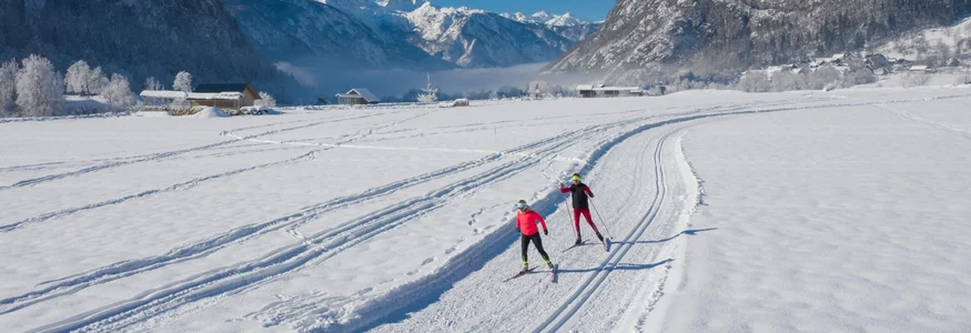 Slow Down in Slowenien © Mojca Odar Zwei Langläufer auf schneebedeckter Loipe vor Bergen bei blauem Himmel