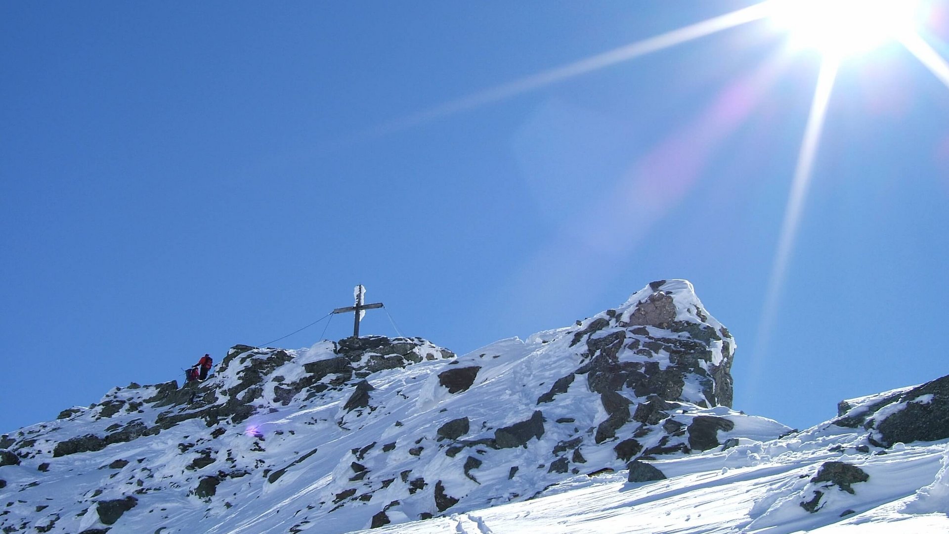Snow-covered mountain peak with summit cross and two hikers under bright sunshine