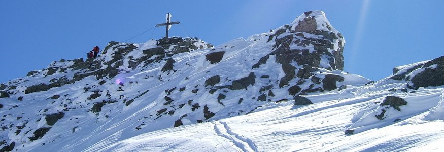 Schneebedeckter Berggipfel mit Gipfelkreuz und zwei Wanderern unter hellem Sonnenschein