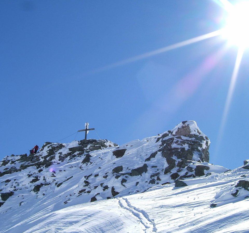 Schneebedeckter Berggipfel mit Gipfelkreuz und zwei Wanderern unter hellem Sonnenschein