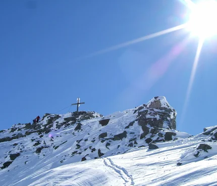 Schneebedeckter Berggipfel mit Gipfelkreuz und zwei Wanderern unter hellem Sonnenschein