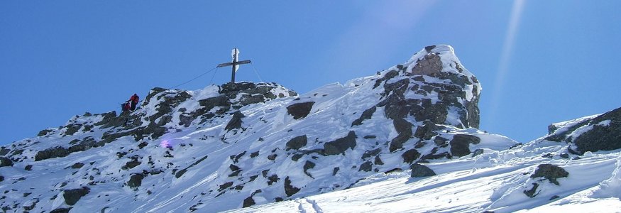 Snow-covered mountain peak with summit cross and two hikers under bright sunshine