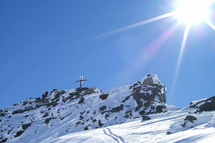 Schneebedeckter Berggipfel mit Gipfelkreuz und zwei Wanderern unter hellem Sonnenschein
