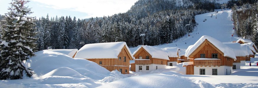Schneebedeckte Holzhäuser vor bewaldeten Bergen und einem Skihang