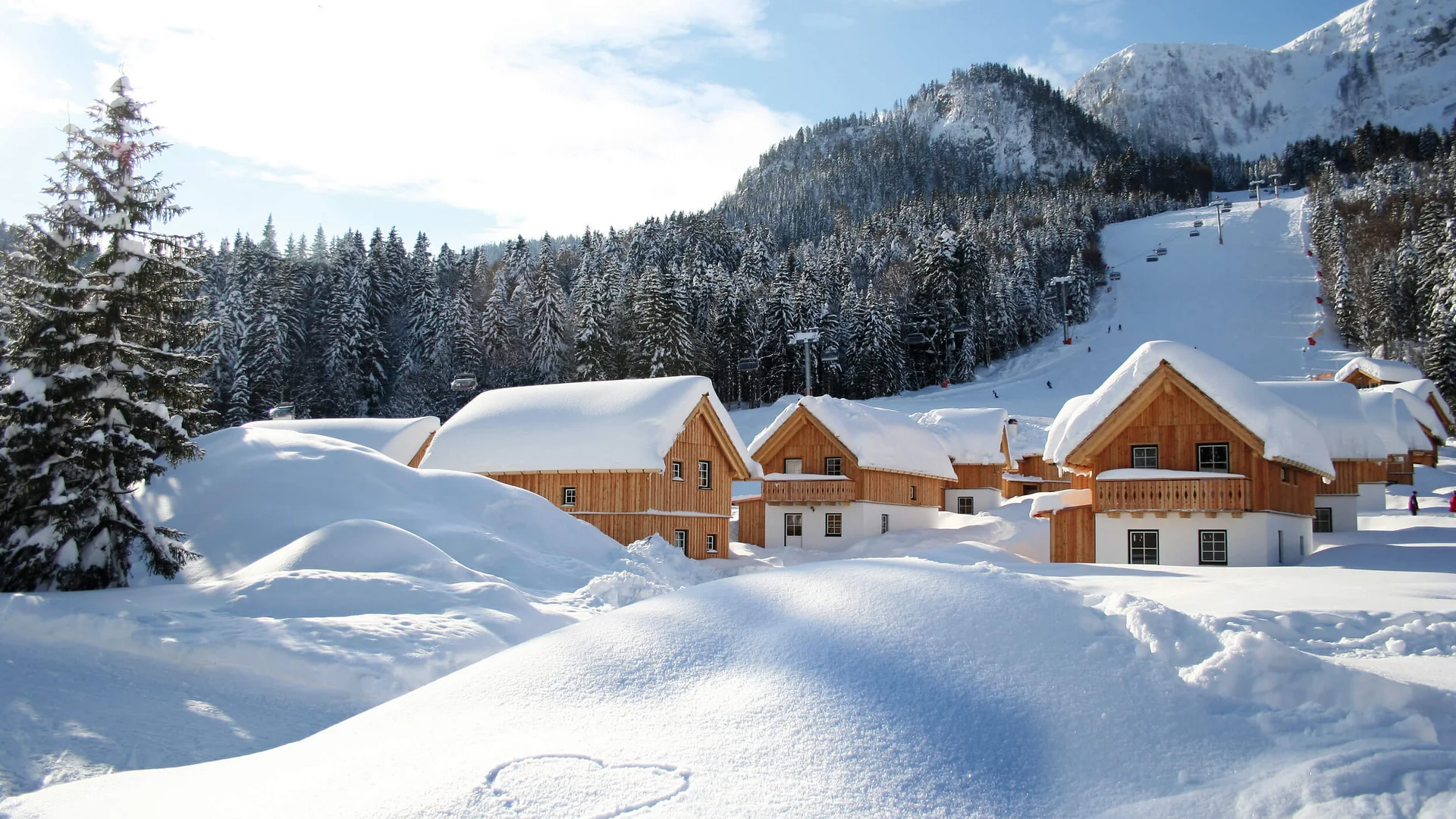 Hüttendorf – Aktiv & Naturresort AlpenParks Hagan Lodge © Gerhard Wolkersdorfer Schneebedeckte Holzhäuser vor bewaldeten Bergen und einem Skihang
