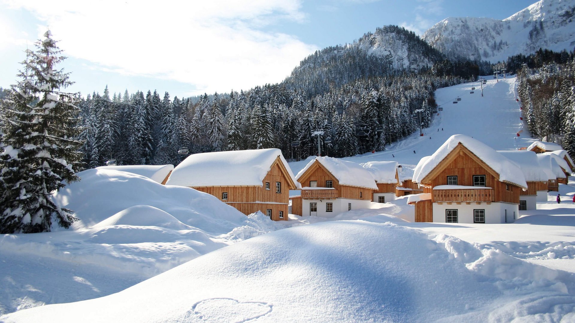 Schneebedeckte Holzhäuser vor bewaldeten Bergen und einem Skihang