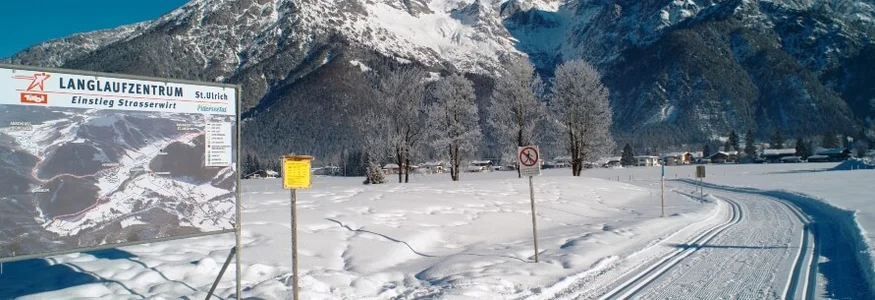 Langlaufzentrum mit gespurter Loipe vor schneebedeckten Bergen und blauem Himmel