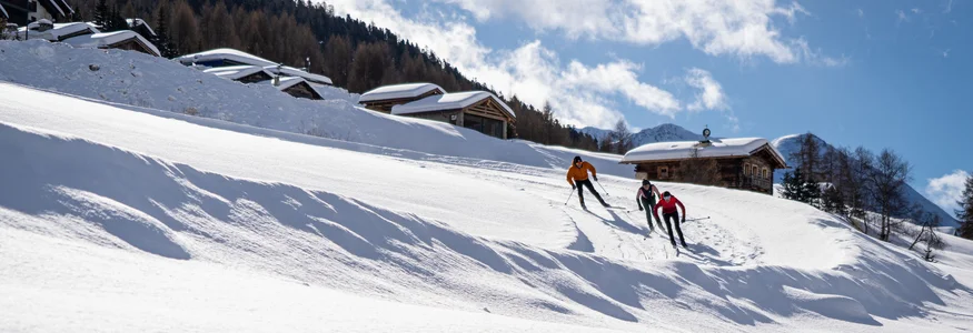 Three skiers skiing past snow-covered cabins in the Alps