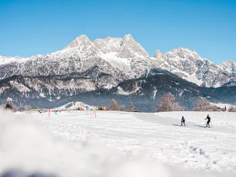 Two cross-country skiers on snowy field with snowy mountains under clear sky