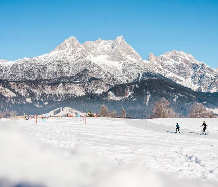 Two cross-country skiers on snowy field with snowy mountains under clear sky