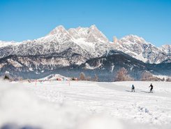Two cross-country skiers on snowy field with snowy mountains under clear sky