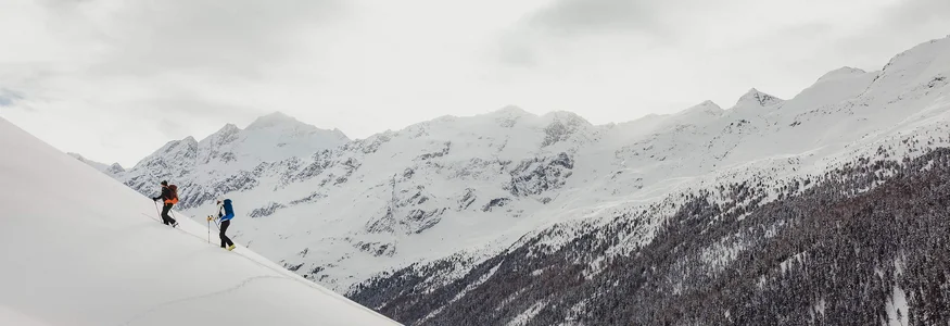 Two skiers climbing a snowy slope with mountain range in the background