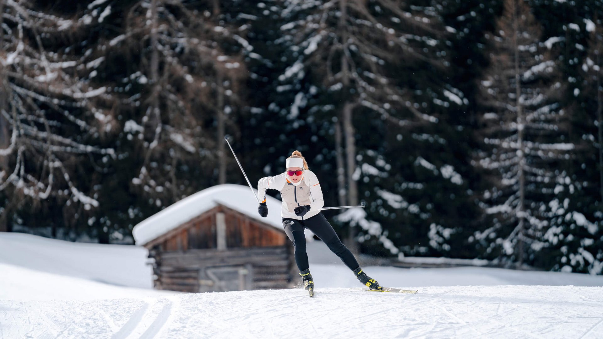 Woman cross-country skiing in front of snowy cabin and forest