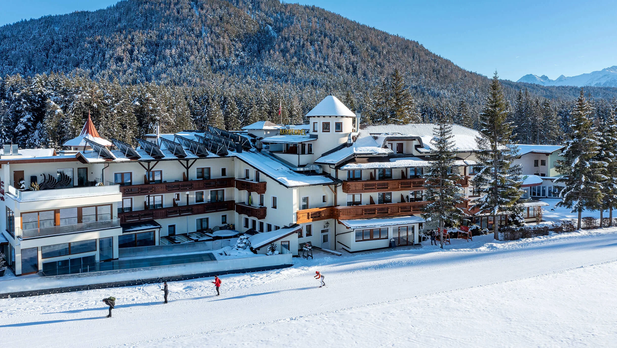 Langlaufgenuss im Karwendel © Hotel Karwendel Bernhard Rödlach BetriebsGmbH Hotelgebäude im Schnee vor bewaldetem Berg unter klarem blauem Himmel