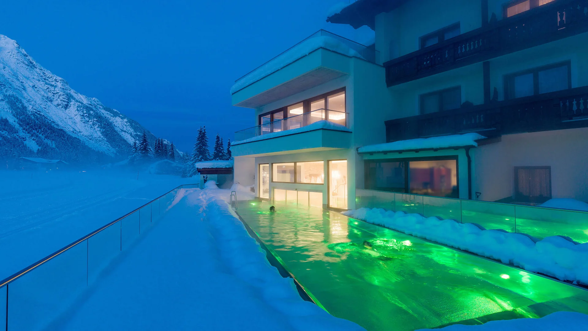 Illuminated outdoor pool beside snowy hotel building in mountains at night