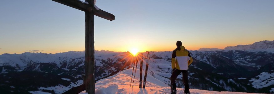 Mann steht neben einem Kreuz auf schneebedecktem Berg bei Sonnenuntergang