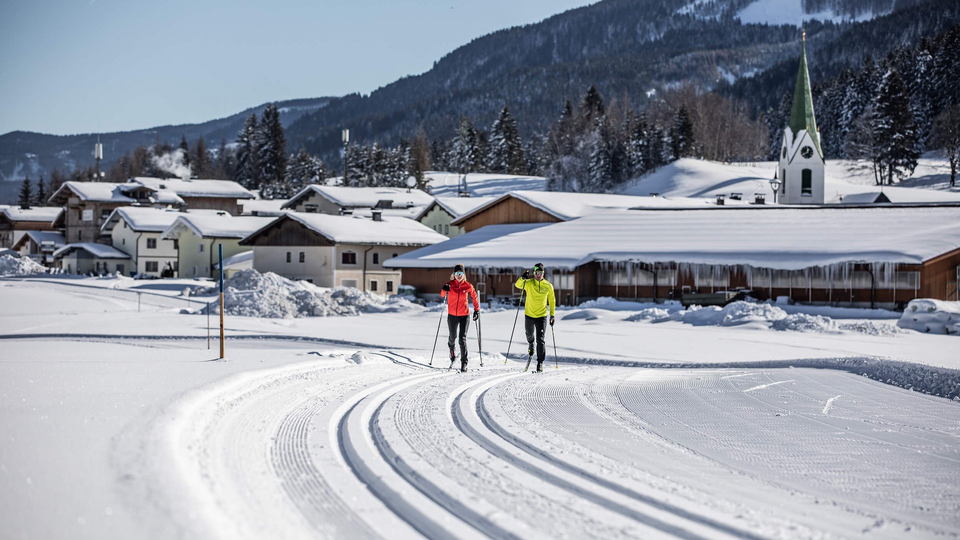 Zwei Langläufer auf gespurteter Loipe vor verschneitem Dorf und Bergen