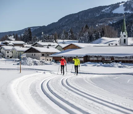 PillerseeTal © Mirja Geh Zwei Langläufer auf gespurteter Loipe vor verschneitem Dorf und Bergen