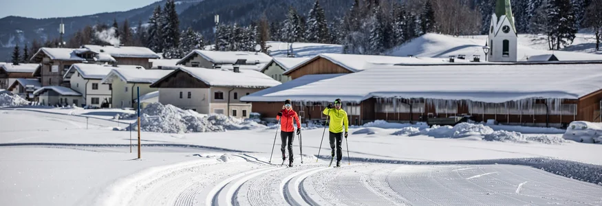 Zwei Langläufer auf gespurteter Loipe vor verschneitem Dorf und Bergen