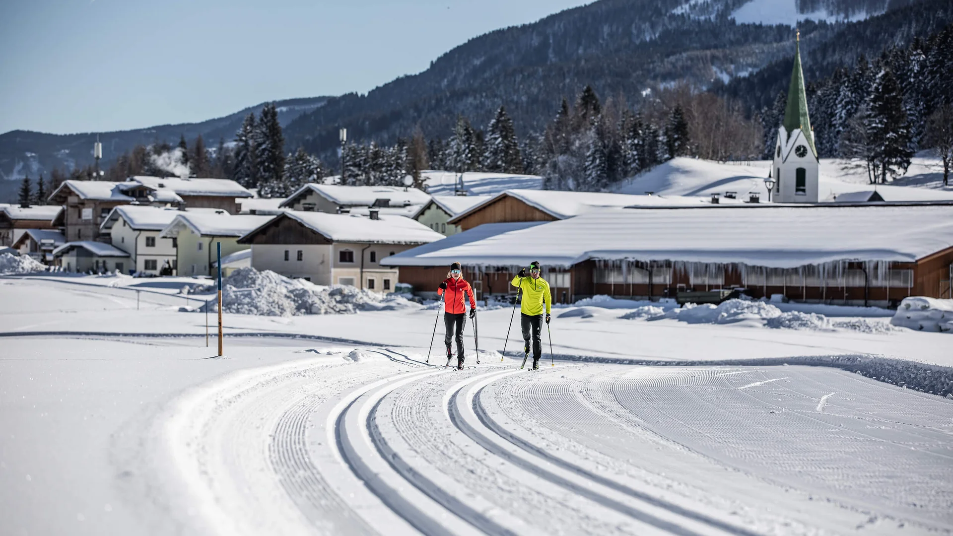 Loipen & Loipenbericht © Mirja Geh Zwei Langläufer auf gespurteter Loipe vor verschneitem Dorf und Bergen