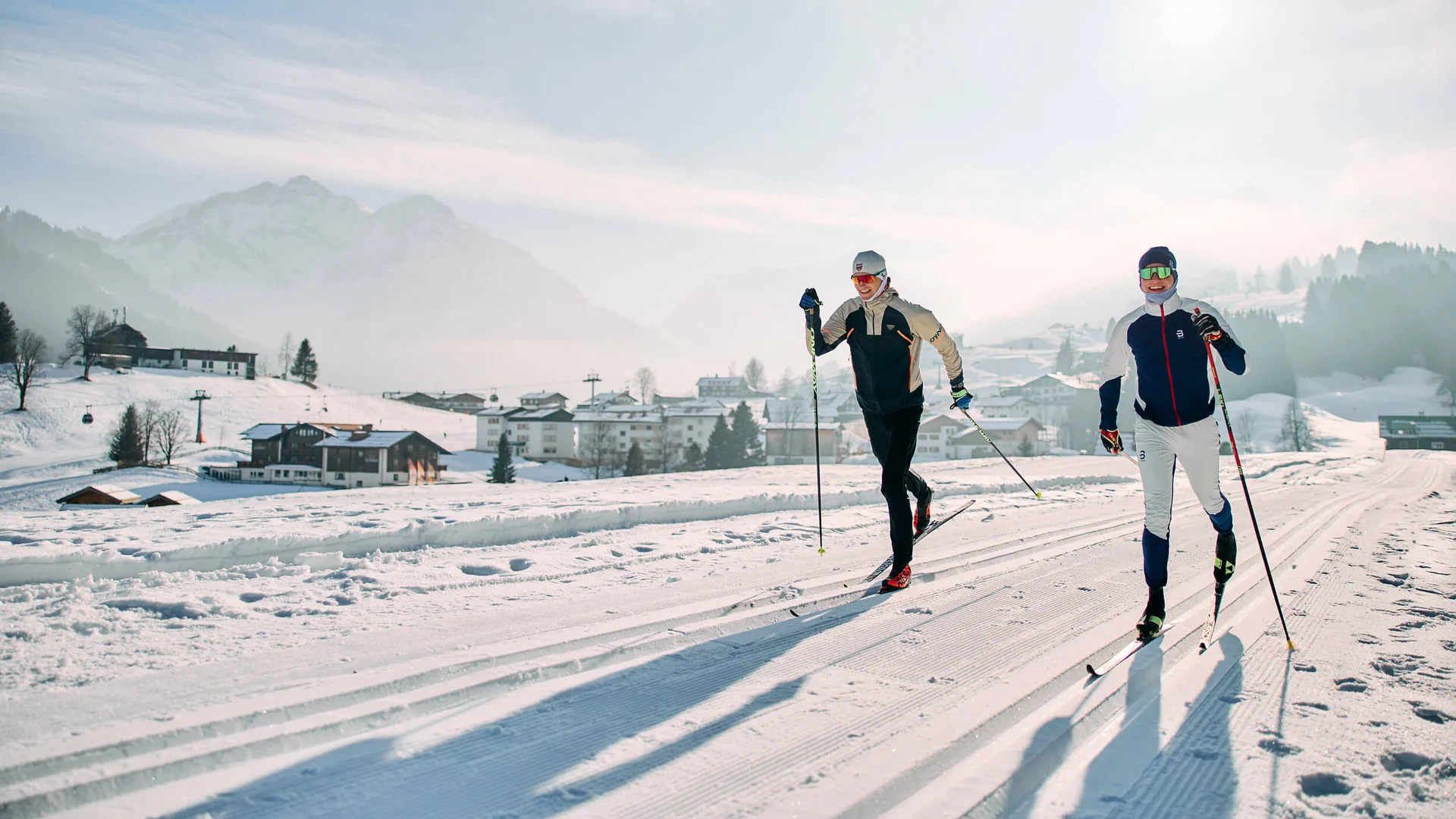 Langlauf Urlaub im Kleinwalsertal © Kleinwalsertal Tourismus - Oliver Farys Zwei Langläufer auf einer verschneiten Loipe vor einem Bergdorf