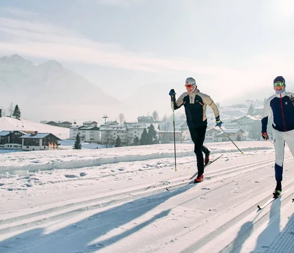 Kleinwalsertal © Kleinwalsertal Tourismus - Oliver Farys Two cross-country skiers on a snowy trail with a mountain village