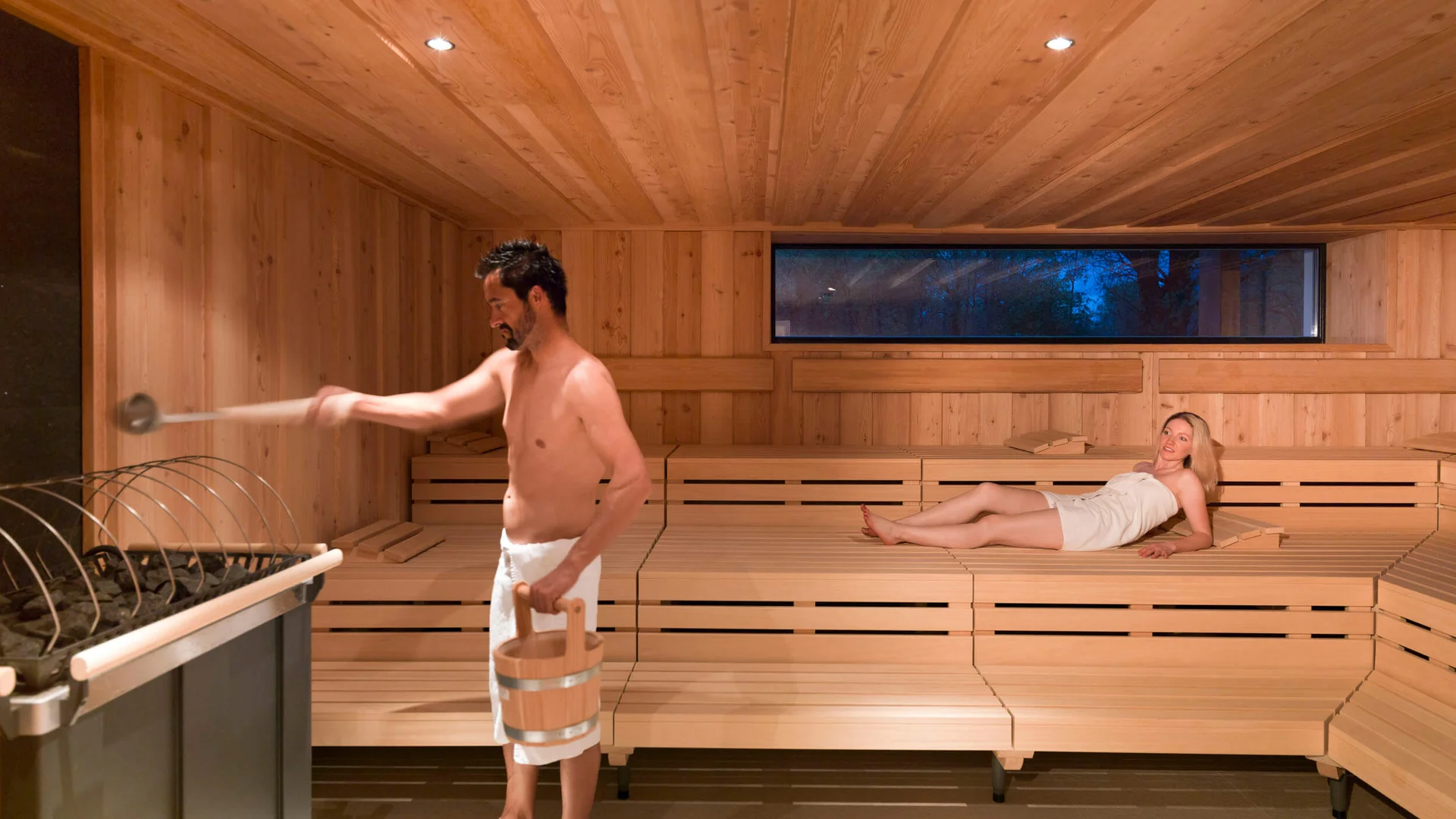 Dolomitengolf Hotel & Spa ****S © Martin Lugger Mann gießt Wasser in die Sauna, Frau liegt entspannt auf der Bank