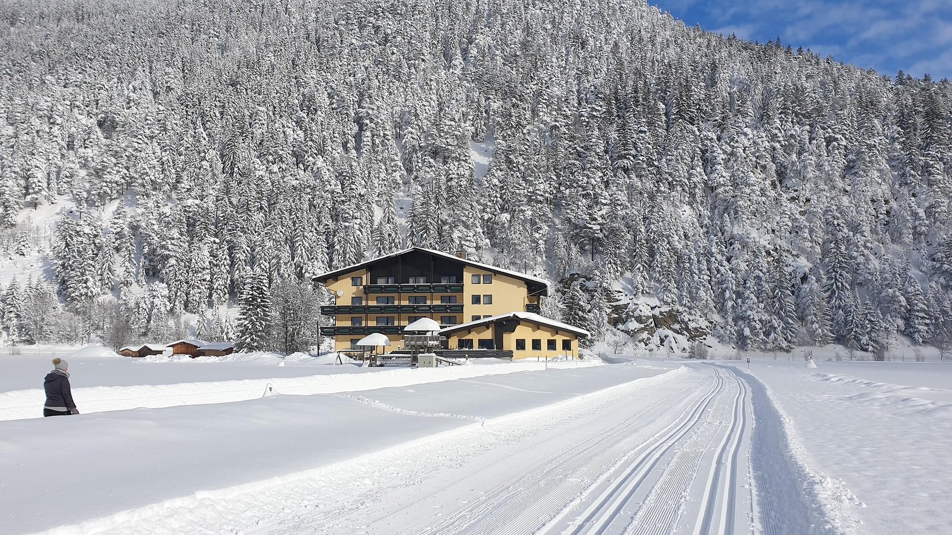 Snow-covered house at the base of a snowy forested hill under blue sky