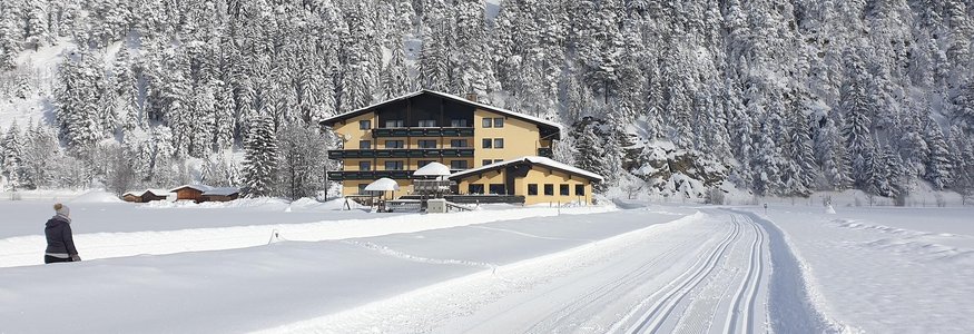 Snow-covered house at the base of a snowy forested hill under blue sky